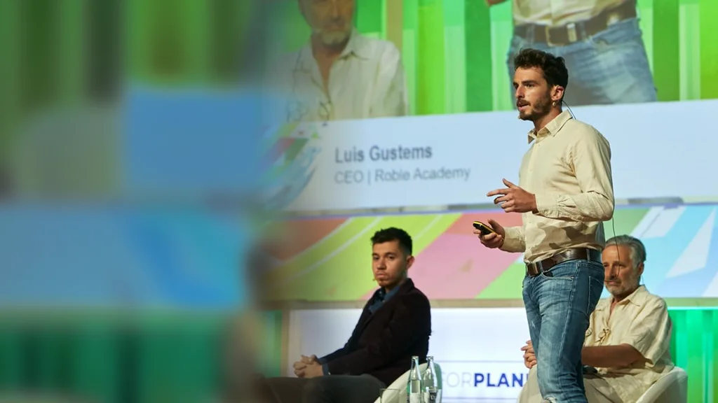 Luis Gustems presentando un evento sobre el escenario, con su nombre en la pantalla del fondo
